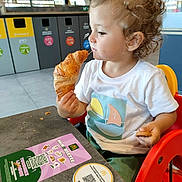 Francis a rejoint le concours — aidez-le/la à gagner de superbes lots ! toddler, child, croissant, cookie, high_chair, curly_hair, white_shirt, table, restaurant, recycling_bins, floor, window, promotion, food, hands, face, side_view, indoor, plastic_chair, meal