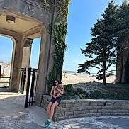 David is registered to the contest to win money with this photo: woman, baby, beach, sand, stone_wall, tree, ivy, sunlight, sky, outdoor, nature, person, holding, casual_clothing, shoes, shorts, sunglasses, greenery, daytime, relaxation