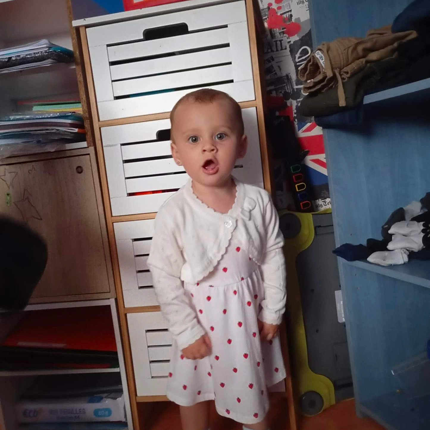 Inaya participe au concours pour gagner de l'argent avec cette photo : cabinet, cardigan, child, clothing, curious, expression, face, floor, furniture, hair, indoor, person, room, shelves, shoes, standing, storage, toddler, white_dress, young_child