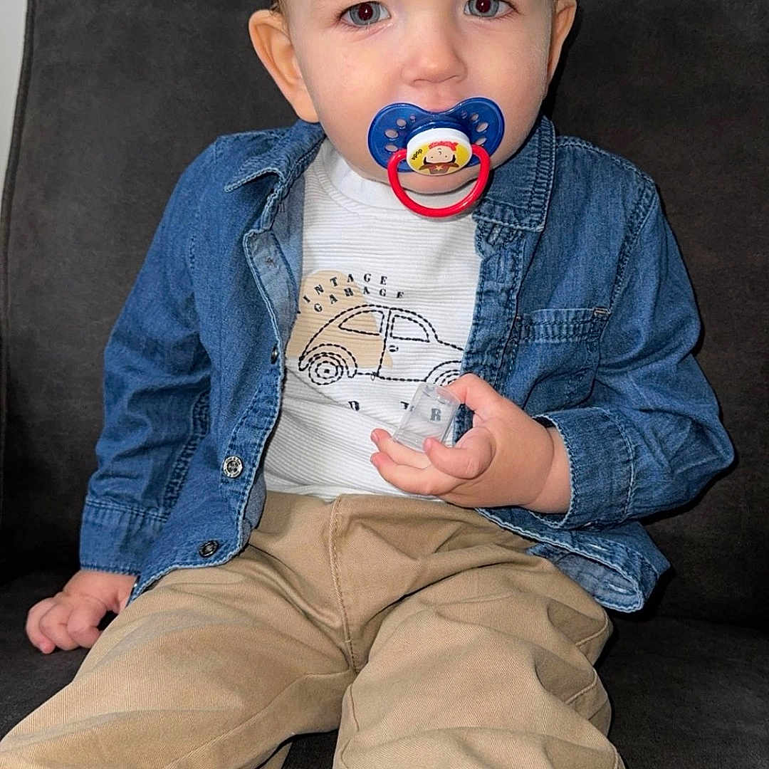Ethan participe au concours pour gagner de l'argent avec cette photo : beige_pants, casual, child, clothing, couch, cute, denim_jacket, expression, face, furniture, hair, indoor, pacifier, person, portrait, sitting, small_object, toddler, white_shoes, young