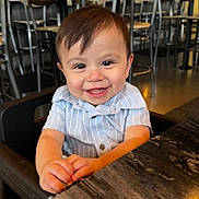 Hudson is registered to the contest to win money with this photo: toddler, child, smiling, happy, high_chair, restaurant, table, clothing, face, person, cute, indoor, baby, hands, seat, wooden_table, short_sleeve, hair, portrait, joyful