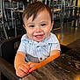 toddler, child, smiling, happy, high_chair, restaurant, table, clothing, face, person, cute, indoor, baby, hands, seat, wooden_table, short_sleeve, hair, portrait, joyful