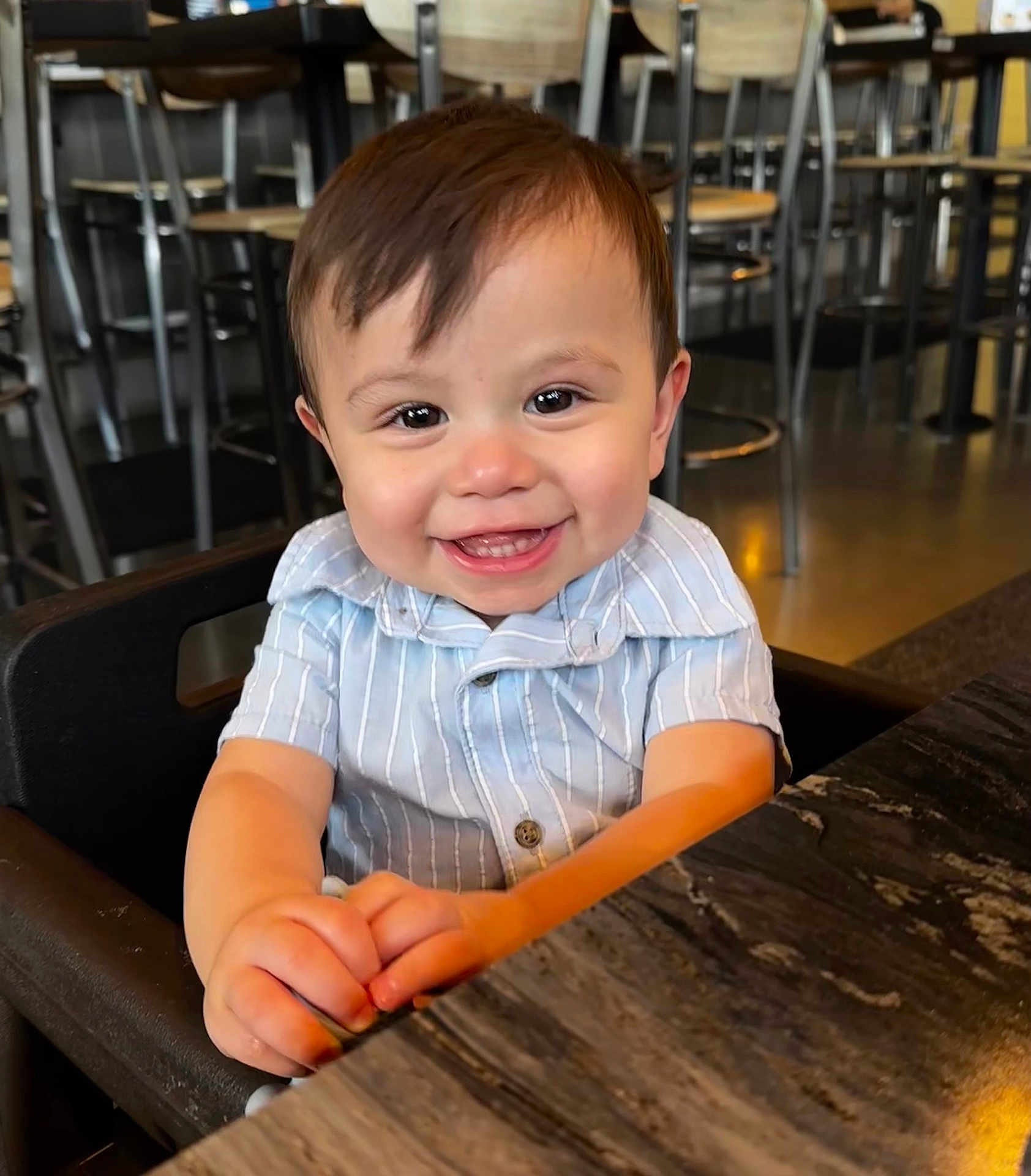 Hudson is registered to the contest to win money with this photo: toddler, child, smiling, happy, high_chair, restaurant, table, clothing, face, person, cute, indoor, baby, hands, seat, wooden_table, short_sleeve, hair, portrait, joyful