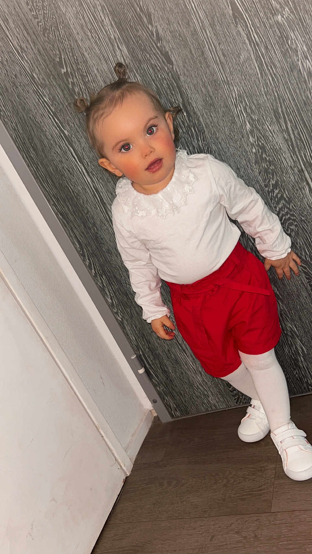 Aïna participe au concours pour gagner de l'argent avec cette photo : child, toddler, portrait, standing, indoor, red_shorts, white_top, white_tights, white_shoes, hair_buns, red_eyes, wooden_wall, door, floor, curious_expression, blush_cheeks, flash_photography, fashion, close_up, cute