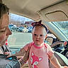 baby, child, woman, car_interior, car_window, parking_lot, sunlight, headband, pink_clothing, fox_design, adult, person, seat, steering_wheel, hand, bracelet, daylight, vehicle, window, outdoor