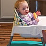 baby, high_chair, toy, red_hair, kitchen, wooden_floor, infant, child, person, seat, straps, purple_clothing, curious, face, indoor, appliance, cabinet, floor, table, home