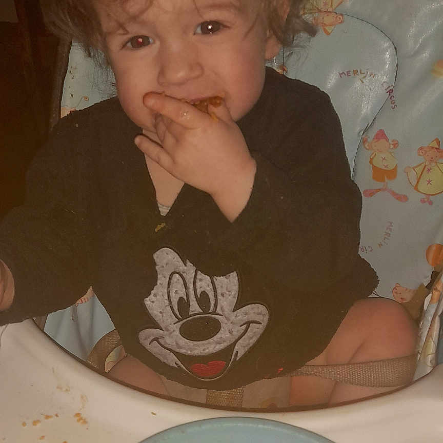 Gabriel a rejoint le concours — aidez-le/la à gagner de superbes lots ! toddler, child, high_chair, mickey_mouse, curly_hair, snack, food, crumbs, plate, happy, messy, indoor, baby, clothing, person, cute, meal, sitting, cartoon, feeding