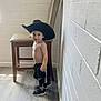 toddler, child, boy, hat, cowboy_hat, shirtless, shorts, sneakers, indoor, floor, chair, wall, smile, standing, looking_at_camera, wood_floor, white_wall, casual, playful, small_person