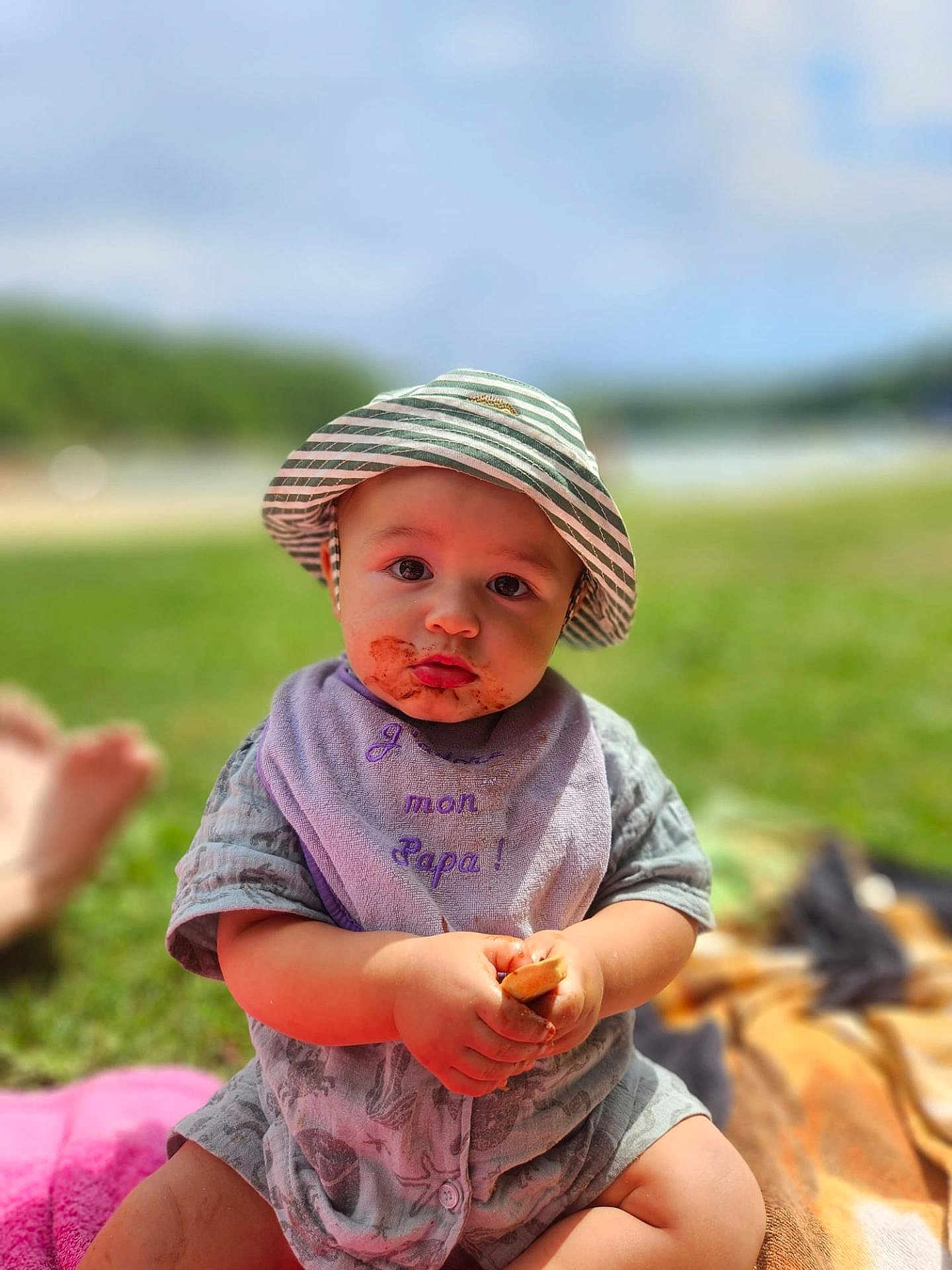 Issam participe au concours pour gagner de l'argent avec cette photo : baby, child, hat, bib, food, messy_face, outdoor, grass, blanket, sunny, summer, sitting, cute, portrait, young_child, clothing, nature, daylight, person, closeup