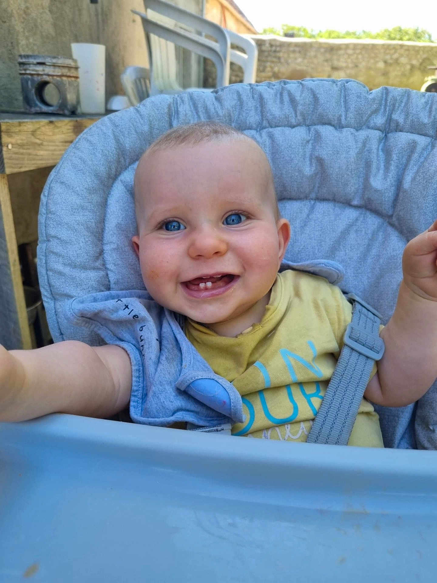 Axel a rejoint le concours — aidez-le/la à gagner de superbes lots ! baby, child, blue_eyes, high_chair, smiling, outdoor, seat_belt, gray_cushion, yellow_tshirt, reaching_hand, face, portrait, happy, infant, person, chair, daylight, casual_clothing, wooden_table, plastic_chair