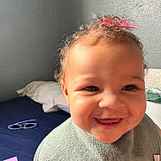 Nariah is registered to the contest to win money with this photo: toddler, child, smiling, curly_hair, pink_bow, green_bib, bed, pillows, glasses, toy_block, red_cup, indoor, happy, face, portrait, playtime, cute, person, young_child, casual