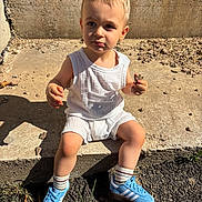 Gabin a rejoint le concours — aidez-le/la à gagner de superbes lots ! bare_arms, blue_sneakers, casual, child, concrete, concrete_wall, curious, daylight, grass, outdoor, rock, shadow, shoes, sitting, socks, step, sunlight, toddler, white_clothing, young_child