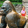Nyomi joined the competition — help win amazing prizes! bronze, child, colorful_clothing, cute, daytime, footwear, gorilla, greenery, happy, nature, outdoor, park, playful, sculpture, sitting, smiling, statue, stone_wall, toddler, yellow_bow