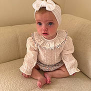 Remi is registered to the contest to win money with this photo: baby, child, infant, girl, headband, bow, blue_eyes, white_clothing, ruffled_collar, sitting, couch, indoor, soft_lighting, cute, portrait, cross_legged, neutral_background, curious_expression, small_feet, long_sleeves
