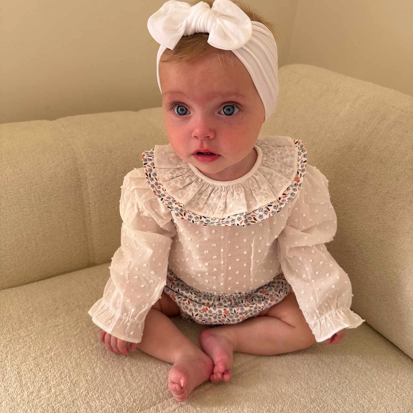 Remi is registered to the contest to win money with this photo: baby, blue_eyes, bow, child, couch, cross_legged, curious_expression, cute, girl, headband, indoor, infant, long_sleeves, neutral_background, portrait, ruffled_collar, sitting, small_feet, soft_lighting, white_clothing