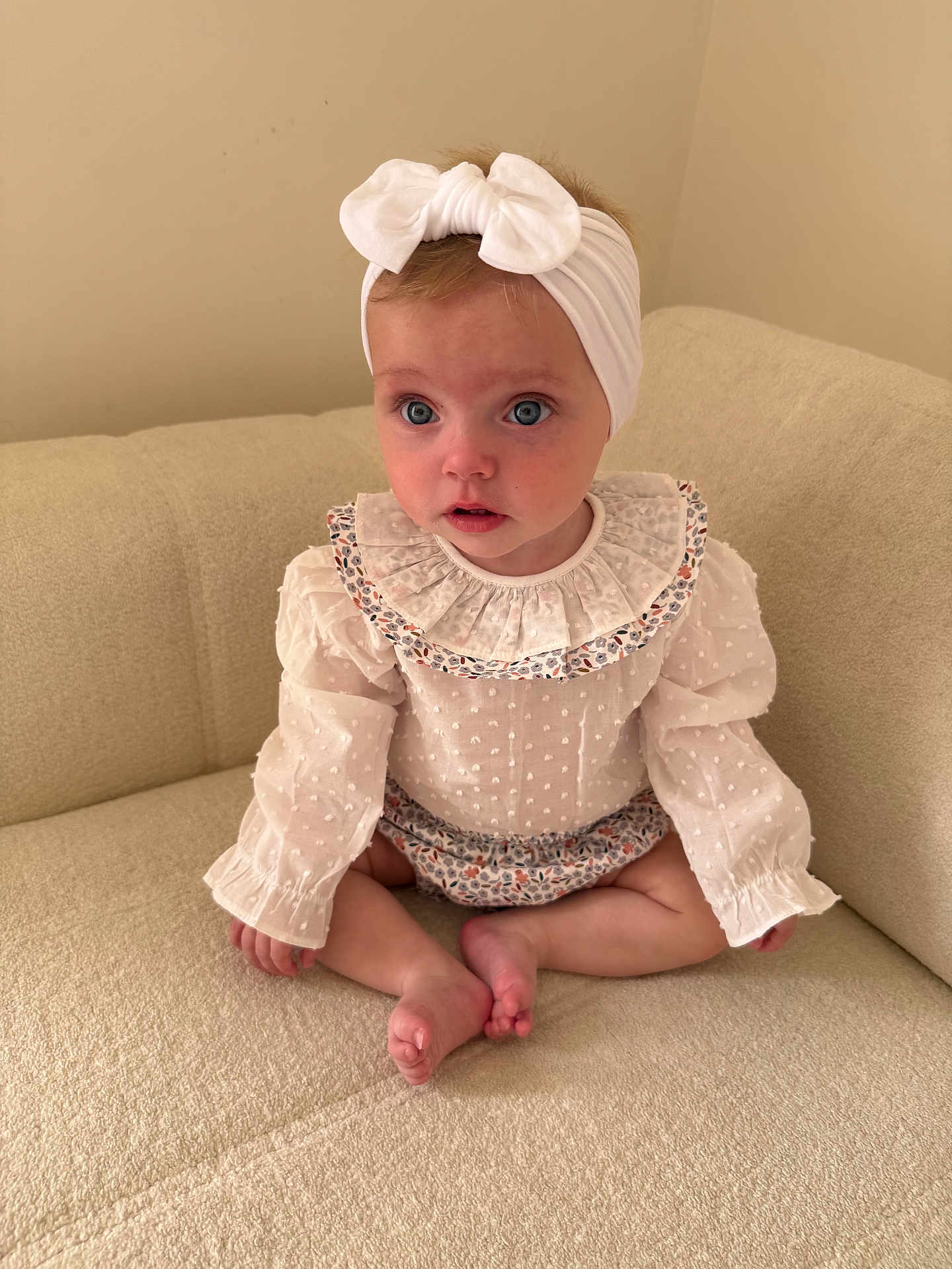 Remi is registered to the contest to win money with this photo: baby, child, infant, girl, headband, bow, blue_eyes, white_clothing, ruffled_collar, sitting, couch, indoor, soft_lighting, cute, portrait, cross_legged, neutral_background, curious_expression, small_feet, long_sleeves