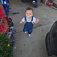 Abel is registered to the contest to win money with this photo: child, toddler, pacifier, denim_overalls, sneakers, concrete_floor, garage, motorcycle, license_plate, tool_cart, extension_cord, plant, yellow_flowers, car_bumper, reflection, standing, looking_up, curious_expression, hose, clutter
