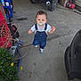 child, toddler, pacifier, denim_overalls, sneakers, concrete_floor, garage, motorcycle, license_plate, tool_cart, extension_cord, plant, yellow_flowers, car_bumper, reflection, standing, looking_up, curious_expression, hose, clutter