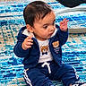 baby, child, infant, sitting, portrait, indoor, rug, carpet, blue_outfit, hoodie, sneakers, bear_design, hands_raised, short_hair, looking_sideways, playful, floor, photograph, smartphone_screenshot, black_border