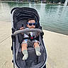 baby, child, stroller, sunglasses, lakeside, lake, fountain, outdoor, park, water, autumn, trees, houses, sidewalk, infant, seated, portrait, socks, gray_stroller, daylight