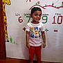child, boy, barefoot, red_pants, white_tshirt, cartoon_characters, giraffe_mural, numbers, educational_wall, indoor, smiling, short_hair, wall_art, colorful, floor, standing, young_child, playful, learning, happy