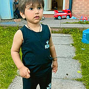 Shivaay is registered to the contest to win money with this photo: child, toddler, outdoor, grass, toy, fire_truck, pathway, black_clothing, sneakers, curious, standing, brick_wall, door, window, bracelet, shorts, sleeveless_shirt, daylight, garden, playtime