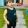 child, toddler, outdoor, grass, toy, fire_truck, pathway, black_clothing, sneakers, curious, standing, brick_wall, door, window, bracelet, shorts, sleeveless_shirt, daylight, garden, playtime