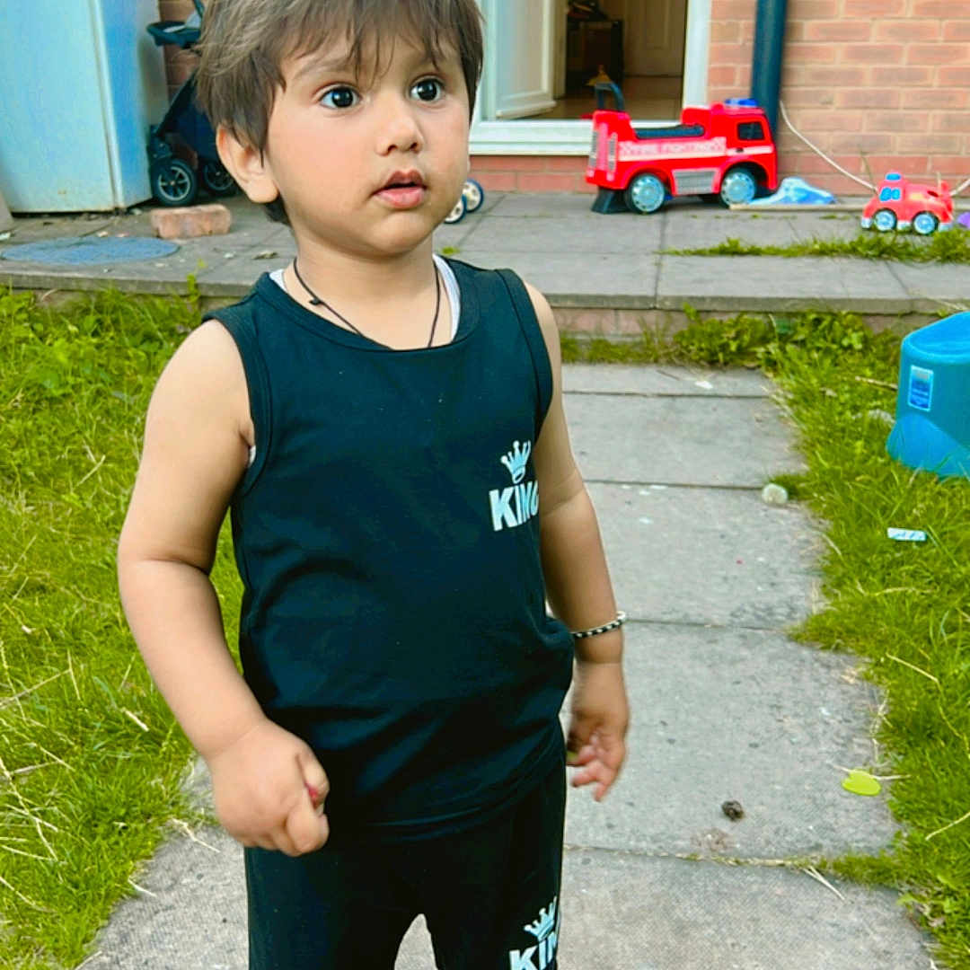 Shivaay is registered to the contest to win money with this photo: black_clothing, bracelet, brick_wall, child, curious, daylight, door, fire_truck, garden, grass, outdoor, pathway, playtime, shorts, sleeveless_shirt, sneakers, standing, toddler, toy, window