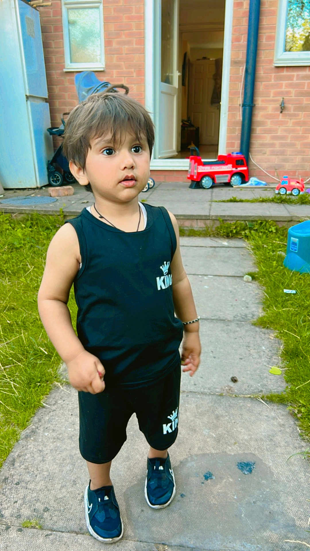 Shivaay is registered to the contest to win money with this photo: child, toddler, outdoor, grass, toy, fire_truck, pathway, black_clothing, sneakers, curious, standing, brick_wall, door, window, bracelet, shorts, sleeveless_shirt, daylight, garden, playtime
