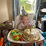 baby, birthday, high_chair, cake, candle, hat, toy, window, curtains, balloons, table, floor, decorations, person, indoor, furniture, plate, food, celebration, child
