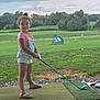 child, girl, golf, driving_range, grass, smile, summer, outdoor, overalls, sandals, golf_club, greenery, sport, fun, playing, nature, happy, young, recreation, activity