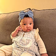 Maryam is registered to the contest to win money with this photo: baby, child, headband, blue_bow, plaid_dress, couch, sofa, indoor, curious, portrait, infant, clothing, seated, skin, person, furniture, cute, expression, hand, cozy