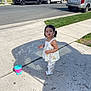 toddler, child, girl, sidewalk, bubble_machine, bubbles, outdoor, sunny, residential_area, plants, grass, vehicles, car, pickup_truck, black_suv, concrete, shadow, summer_clothing, pigtails, playtime