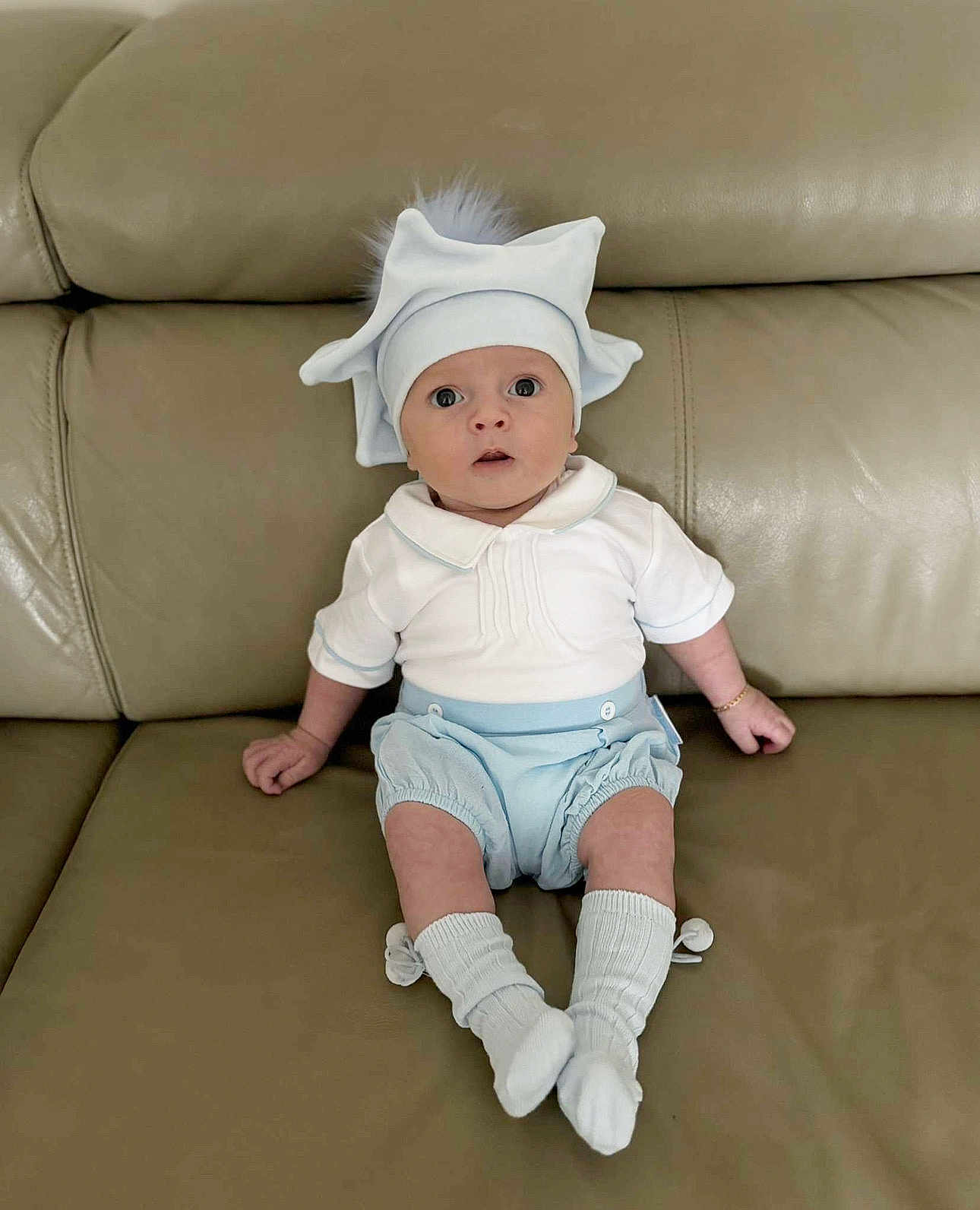 Reaves is registered to the contest to win money with this photo: baby, infant, hat, white_clothing, blue_clothing, socks, couch, leather, sitting, wide_eyes, cute, adorable, child, indoor, portrait, expression, pom_pom, casual, furniture, young