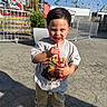 amusement_park, boy, carnival, casual_clothing, child, daytime, drink, fence, ferris_wheel, fun, outdoor, pavement, plant, portrait, smiling, sneakers, straw, sunny, tshirt, young