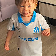 Ethan participe au concours pour gagner de l'argent avec cette photo : toddler, child, smiling, indoor, soccer_uniform, white_clothing, blue_clothing, toy, floor, sofa, table, person, legs, sock, happy, playful, home, young_child, standing, casual