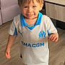 toddler, child, smiling, indoor, soccer_uniform, white_clothing, blue_clothing, toy, floor, sofa, table, person, legs, sock, happy, playful, home, young_child, standing, casual