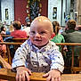 baby, child, smiling, indoor, church, wooden_pew, people, audience, patterned_shirt, hands, face, blond_hair, religious_art, seated, background, casual_clothing, crowd, portrait, happy, event