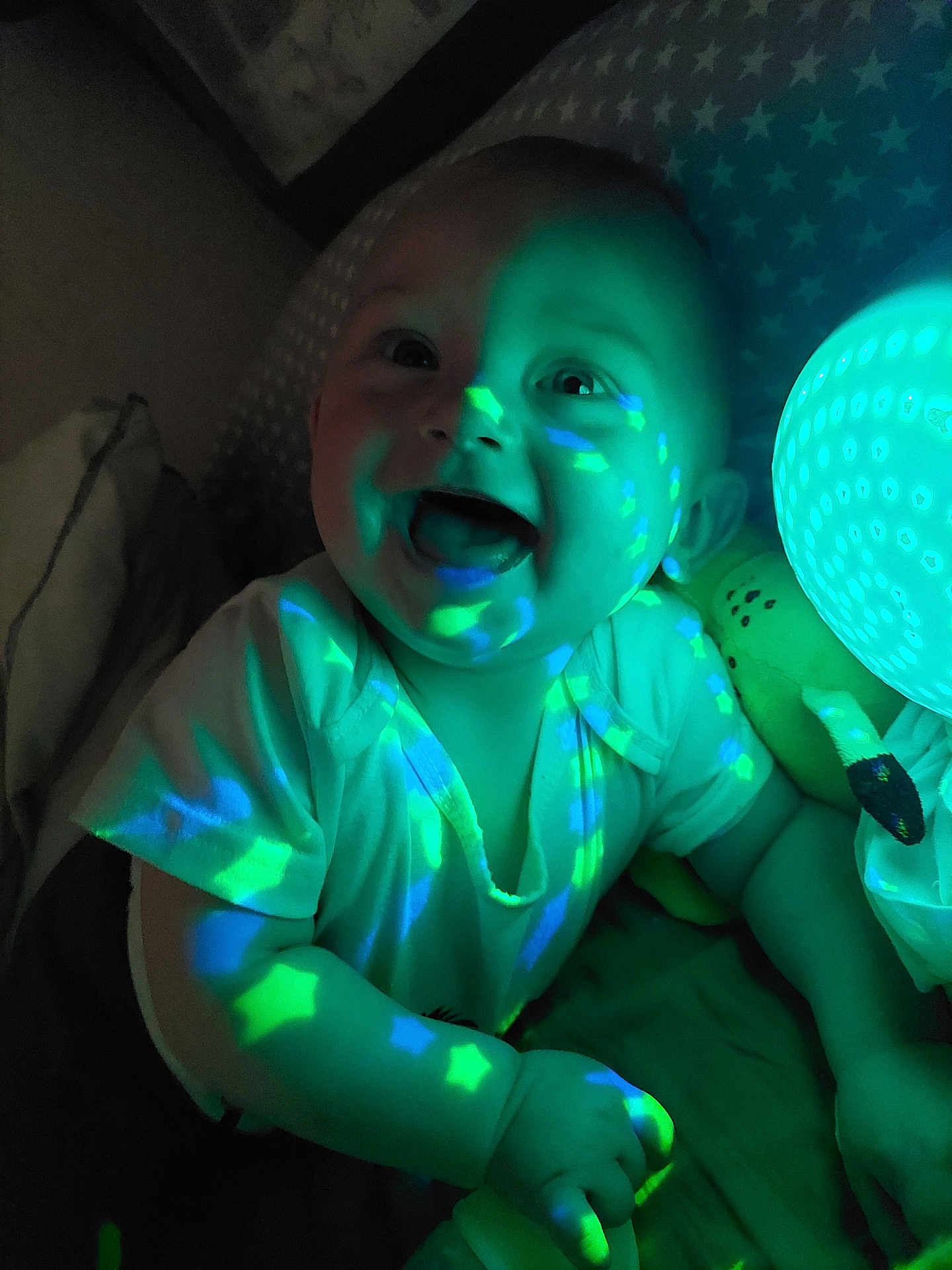 Eddy participe au concours pour gagner de l'argent avec cette photo : baby, child, glowing_light, star_pattern, blue_light, green_light, plush_toy, smiling, happy, indoor, nighttime, projection, cute, infant, glowing_orb, short_sleeve_shirt, face, hand, bed, cozy