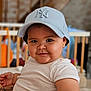 apparel, baby, baseball_cap, blurred_background, cap, child, chubby_cheeks, closeup, cute, face, happy, headwear, indoor, infant, person, play_area, portrait, smile, white_shirt, young