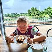 Lazarus is registered to the contest to win money with this photo: boy, cafe, cake, casual_clothing, child, coffee, cup, drink, food, frosting, greenery, nuts, outdoor, plate, sky, smile, table, tree, window, wood