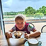 boy, cafe, cake, casual_clothing, child, coffee, cup, drink, food, frosting, greenery, nuts, outdoor, plate, sky, smile, table, tree, window, wood
