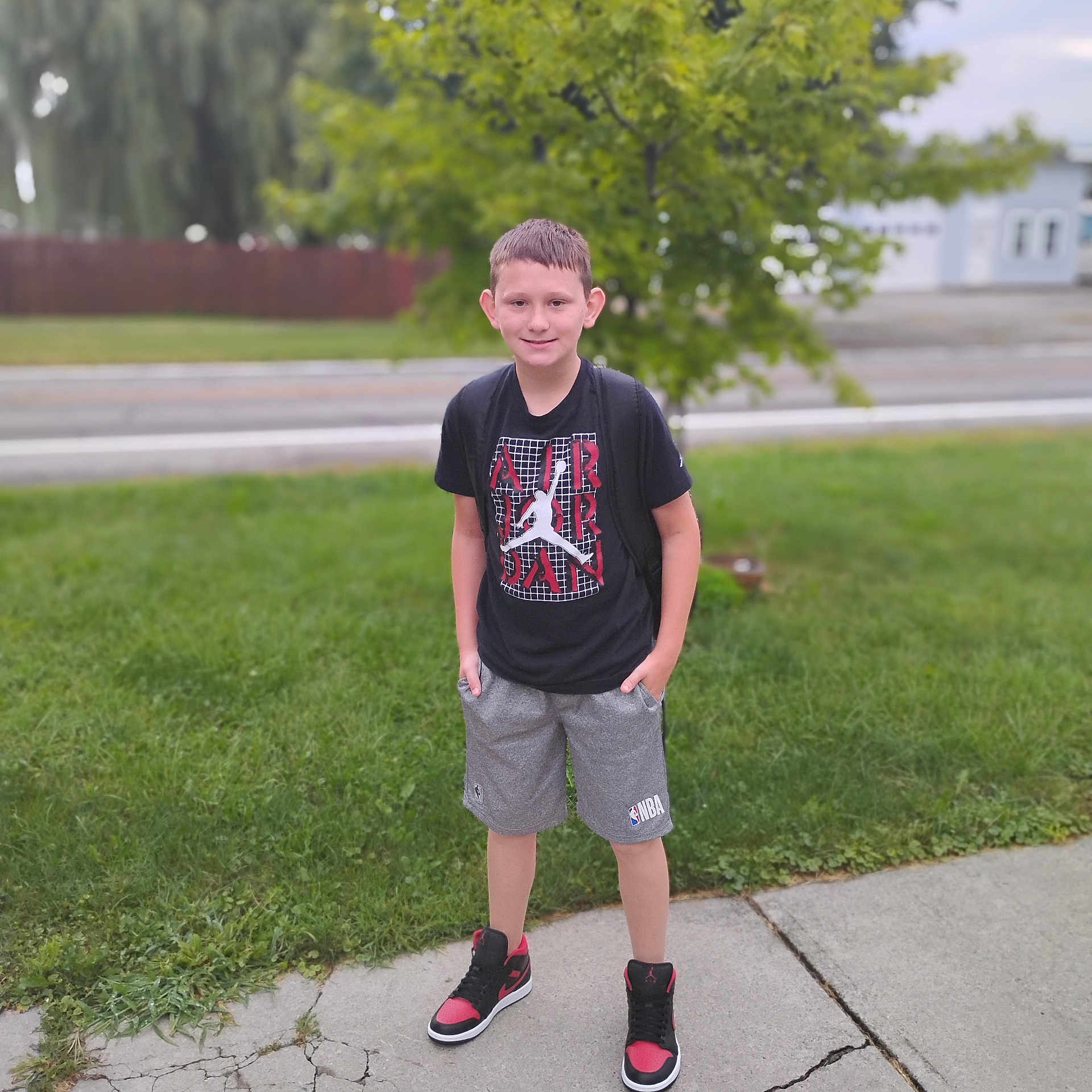 Ryder is registered to the contest to win money with this photo: boy, child, smile, standing, sidewalk, grass, tree, street, house, backpack, casual_clothing, tshirt, shorts, sneakers, outdoor, suburban, summer, young, person, portrait