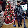 bookshelf, bow_tie, boy, celebration, child, christmas_tree, curtain, decorations, festive, flooring, furniture, holiday, indoor, lights, ornaments, paper_chain, plaid_shirt, smile, television, wood_floor
