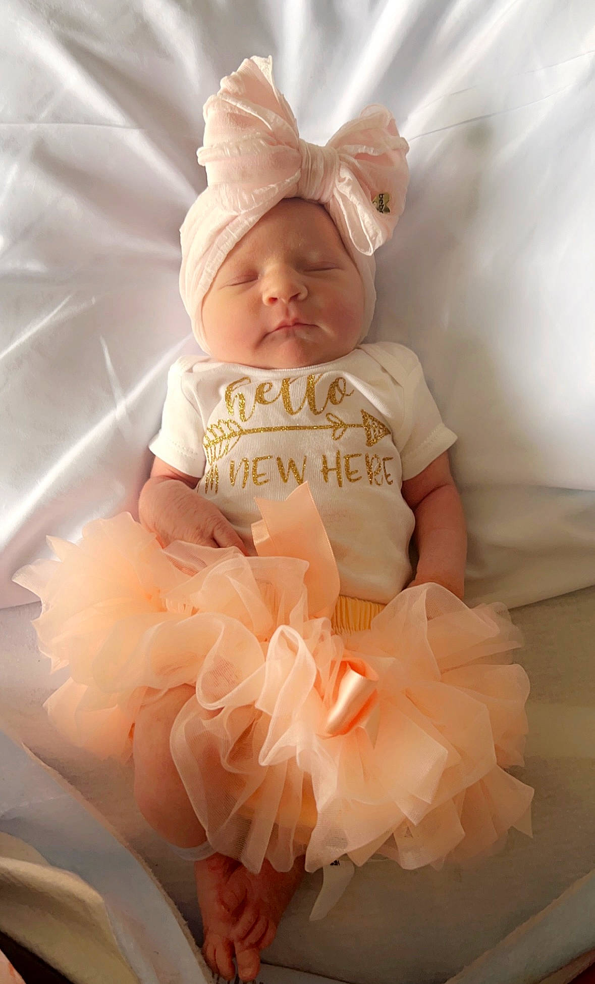 Nora is registered to the contest to win money with this photo: baby, baby_toddler_clothing, clothing, dress, embellishment, event, face, fashion_accessory, hair_accessory, happy, headgear, headpiece, headwear, outerwear, pattern, peach, person, pink, ruffle, sleeve