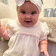 Kinsley is registered to the contest to win money with this photo: baby, child, infant, portrait, smiling, happy, bow, headband, dress, pink_clothing, sitting, couch, indoor, home, phone_screen, call_interface, smartphone, ios, cozy, face