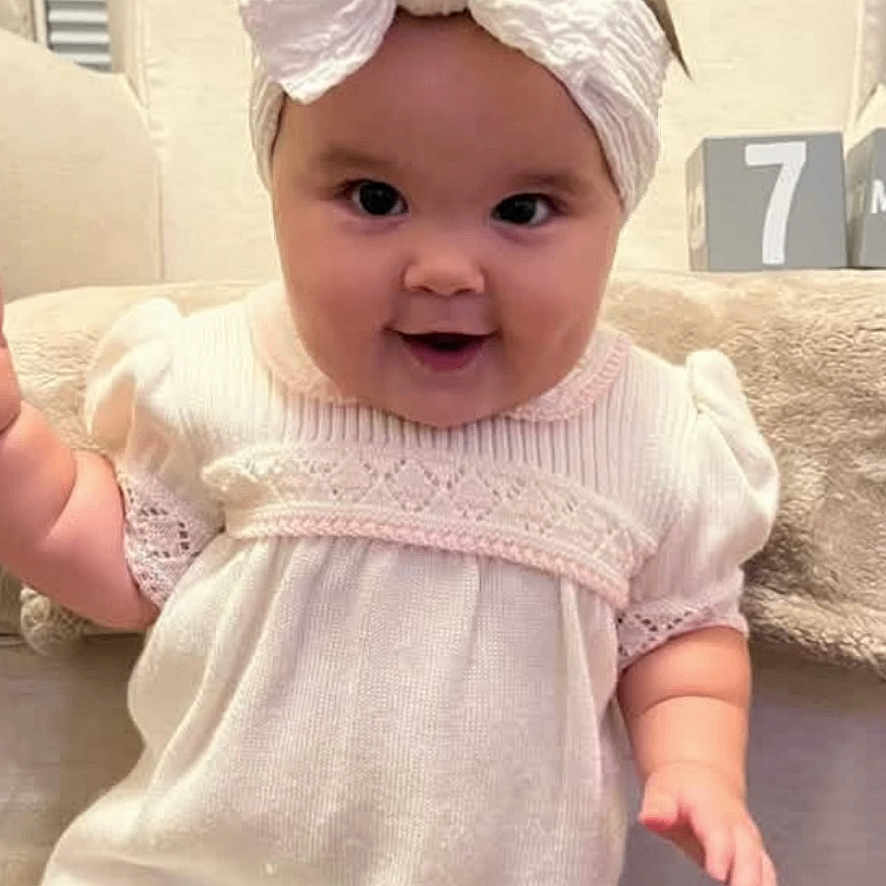 Kinsley is registered to the contest to win money with this photo: baby, child, infant, portrait, smiling, happy, bow, headband, dress, pink_clothing, sitting, couch, indoor, home, phone_screen, call_interface, smartphone, ios, cozy, face