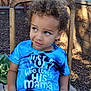 child, boy, curly_hair, blue_shirt, text_on_shirt, wooden_chair, garden, flowers, fence, outdoor, sitting, casual_clothing, natural_light, portrait, young_child, expression, plant, nature, daylight, relaxed