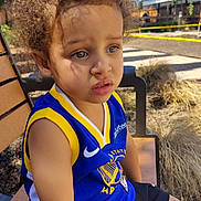 Israel is registered to the contest to win money with this photo: child, toddler, curly_hair, basketball_jersey, blue_jersey, yellow_trim, outdoor, bench, sitting, sunny, daylight, expression, portrait, casual_clothing, shorts, young, person, nature, trees, fence
