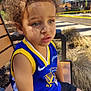 child, toddler, curly_hair, basketball_jersey, blue_jersey, yellow_trim, outdoor, bench, sitting, sunny, daylight, expression, portrait, casual_clothing, shorts, young, person, nature, trees, fence